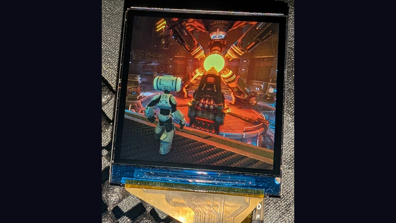 A close-up photo of the small square prototype screen showing a robot facing a glowing reactor, with its ribbon cable below